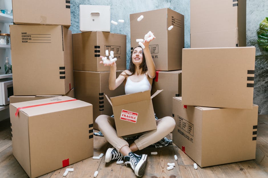A person is sitting on a wooden floor next to a large open cardboard box labeled 'BOOKS' with black handwritten text. Several smaller stacks of books, tied with twine, are placed in front of the box. 