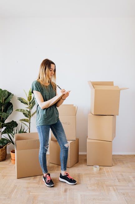 A young male mover with shoulder-length light brown hair, wearing a dark blue coverall and a patterned headband, stands inside a property surrounded by packed moving boxes and packaging materials. He 