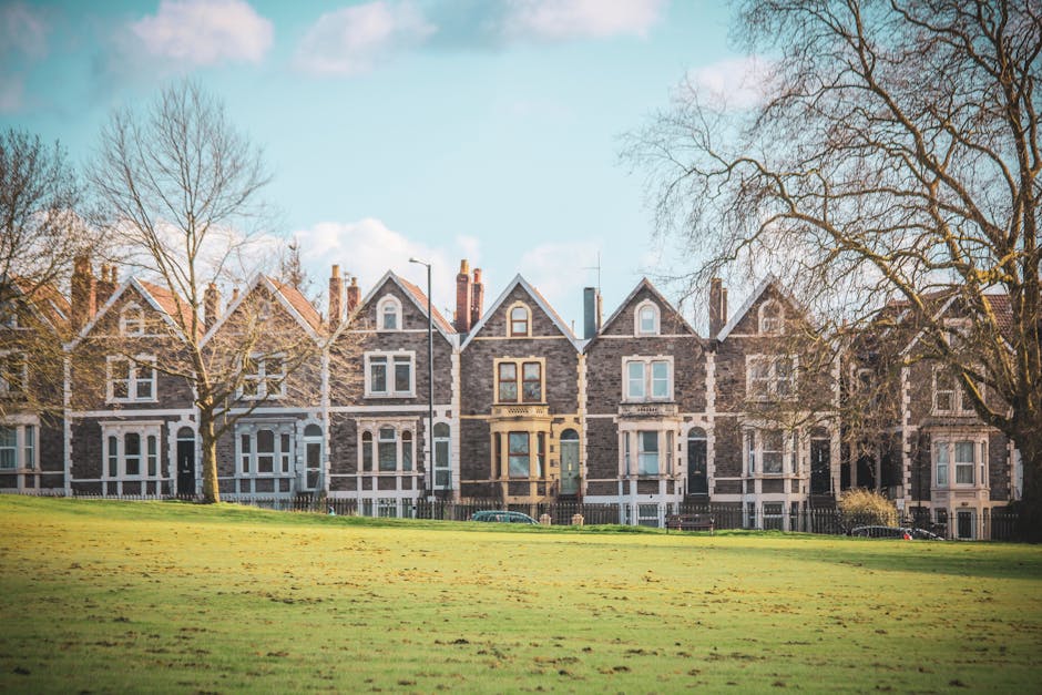 A row of Victorian-style terrace houses constructed from grey stone with white-framed windows and decorative mouldings, set along a tree-lined street under a bright blue sky with scattered clouds. Bar
